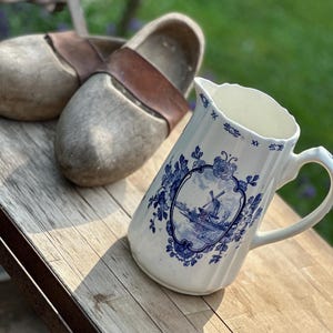 Può includere: Un paio di zoccoli di legno con cinturini in pelle marrone e una brocca bianca con un disegno di mulino a vento blu e bianco. La brocca ha un manico e un bordo smerlato. Gli oggetti sono su una superficie di legno.