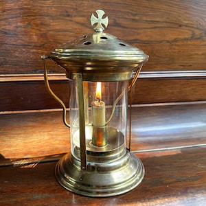 Puede incluir: Una linterna de latón con una vela encendida en su interior. La linterna tiene un cilindro de vidrio transparente, una parte superior en forma de cruz y un asa. La llama de la vela es visible, emitiendo un brillo cálido. La linterna está sobre una superficie de madera.
