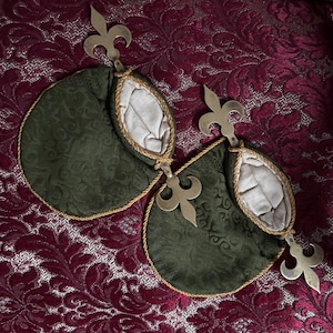 May include: Two dark green fabric pouches with gold trim and silver fleur-de-lis accents. The pouches are connected and have a light-colored interior. The background is a burgundy fabric with a floral pattern.