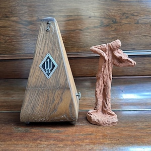Könnte beinhalten: Ein hölzernes Metronom mit einem silbernen Emblem und eine kleine Terrakotta-Skulptur einer Person, die Geige spielt. Das Metronom hat eine Pyramidenform und eine dunkle Holzmaserung. Die Skulptur ist rotbraun.