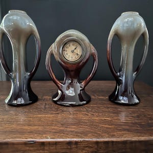 May include: Three ceramic decorative items. Two vases with a tall, open design and a clock in the center. The vases and clock have a dark brown base that fades to a lighter brown and gray at the top. The clock has a cream-colored face.