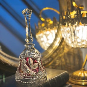 May include: A clear crystal bell with a long handle. The bell has a red interior with a bird and leaf design. The bell is on a stack of books, with a gold mirror and lamp in the background.