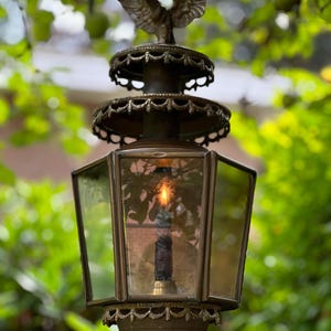Op de afbeelding: Een antieke bronzen buitenlantaarn met een decoratieve adelaar. De lantaarn heeft een glazen behuizing met een brandende kaars. De achtergrond is wazig groen gebladerte.