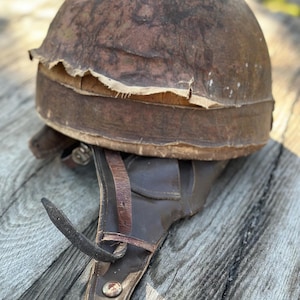 Può includere: Un casco antico in pelle marrone con un aspetto usurato. Il casco ha una parte superiore arrotondata e un cinturino in pelle con rivetti metallici. La pelle invecchiata mostra segni di usura, suggerendo la sua età. Il casco è probabilmente dei primi del XX secolo.