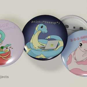 Può includere: Tre spille rotonde a forma di bottone che raffigurano serpenti di cartoni animati con diversi design. La prima spilla ha un serpente blu chiaro in una ciotola rossa di noodles con il testo "DANGER NOOPLE". La seconda spilla ha un serpente blu chiaro e giallo con un laptop e il testo "print("Ssssup")". La terza spilla ha un serpente rosa con il testo "g-s-s-senpai".