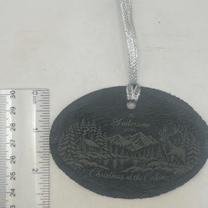 May include: An oval slate ornament with a silver ribbon, featuring an engraved mountain cabin scene, a deer, and the text "The Andersons 2026 Christmas at the Cabin". A ruler shows the ornament is approximately 7.6 cm tall.