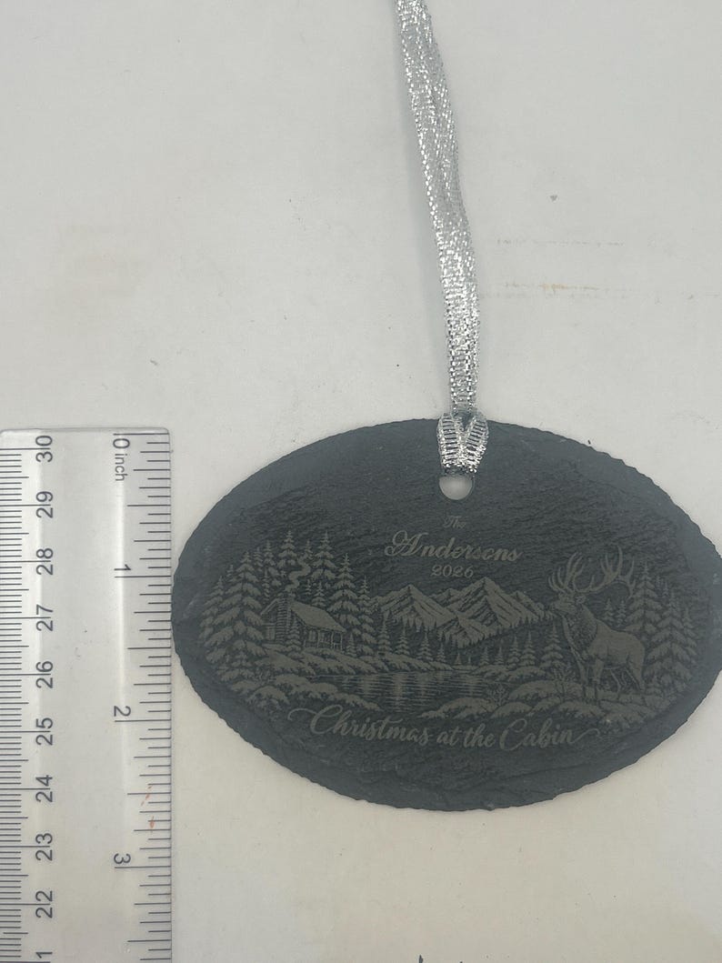 May include: An oval slate ornament with a silver ribbon, featuring an engraved mountain cabin scene, a deer, and the text "The Andersons 2026 Christmas at the Cabin". A ruler shows the ornament is approximately 7.6 cm tall.