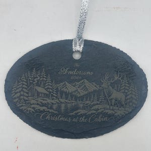 May include: An oval slate ornament with a silver ribbon featuring an engraved winter scene of a cabin, mountains, and a deer. The text reads The Andersons 2026 and Christmas at the Cabin. The dark grey stone has a rough, natural edge texture.