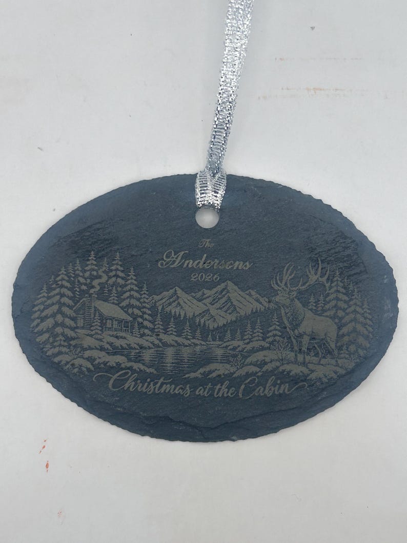 May include: An oval slate ornament with a silver ribbon featuring an engraved winter scene of a cabin, mountains, and a deer. The text reads The Andersons 2026 and Christmas at the Cabin. The dark grey stone has a rough, natural edge texture.
