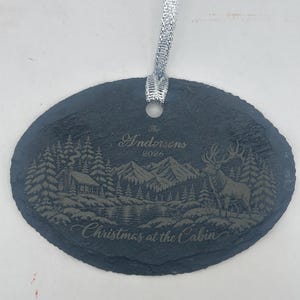 May include: An oval slate ornament with a silver ribbon featuring an engraved mountain landscape, a cabin, a deer, and the text The Andersens 2026 Christmas at the Cabin. The dark grey stone has a rough, natural edge and a smooth, flat surface.