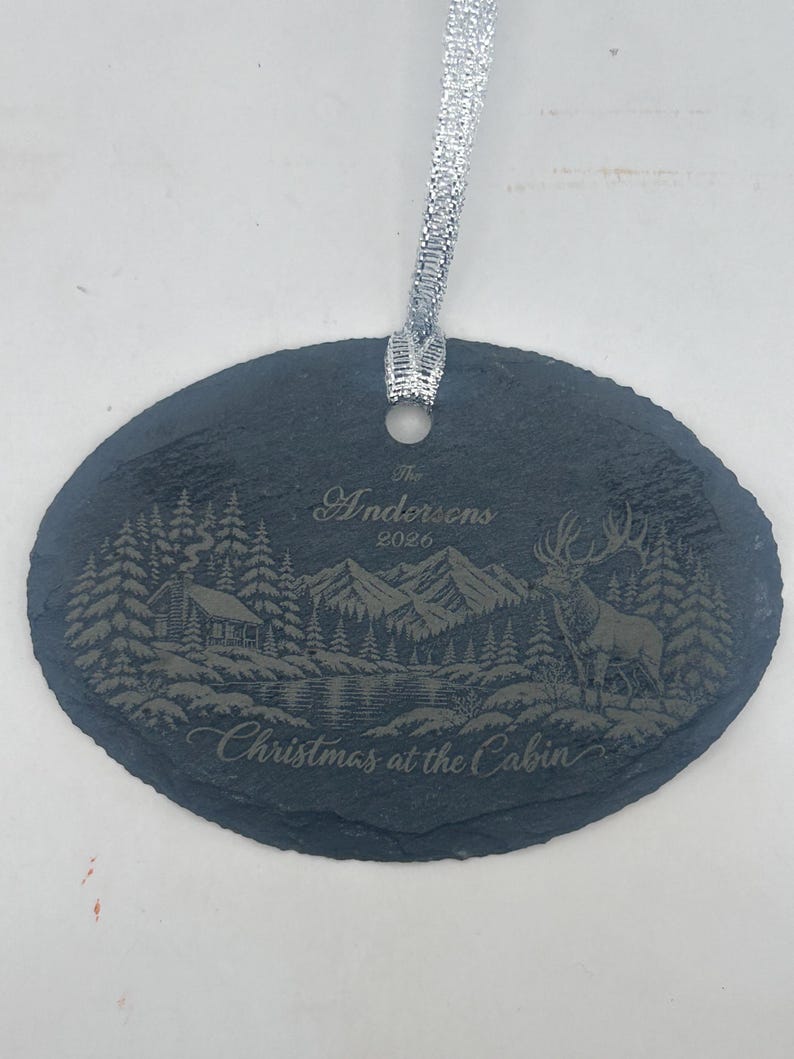 May include: An oval slate ornament with a silver ribbon featuring an engraved mountain landscape, a cabin, a deer, and the text The Andersens 2026 Christmas at the Cabin. The dark grey stone has a rough, natural edge and a smooth, flat surface.