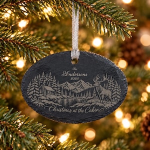 May include: An oval slate Christmas ornament featuring an engraved mountain landscape with a cabin, a lake, and a deer. The text reads The Andersons 2026 and Christmas at the Cabin. It hangs from a silver ribbon against a blurred evergreen tree.