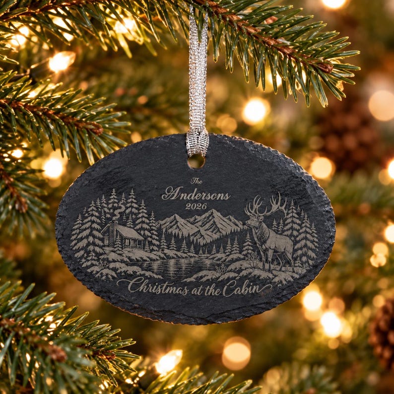May include: An oval slate Christmas ornament featuring an engraved mountain landscape with a cabin, a lake, and a deer. The text reads The Andersons 2026 and Christmas at the Cabin. It hangs from a silver ribbon against a blurred evergreen tree.