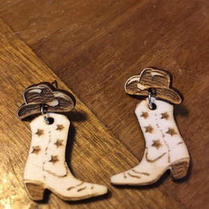 May include: A pair of wooden earrings shaped like cowboy boots with a star pattern and a cowboy hat on top. The earrings are a light brown color.