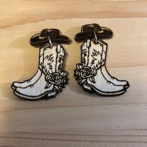 May include: A pair of wooden earrings featuring a cowboy boot design with a flower detail. Each boot has a black cowboy hat on top.
