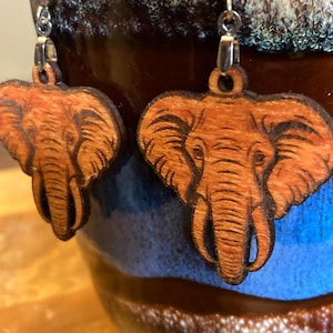 May include: A pair of wooden earrings shaped like elephants. The earrings are a light brown colour and have a detailed design. The earrings are hanging from silver hooks.