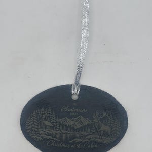 May include: An oval-shaped black slate ornament featuring an engraved mountain landscape with a cabin, a deer, and pine trees. The text reads The Andersons 2026 and Christmas at the Cabin. A silver ribbon is attached through a hole at the top.