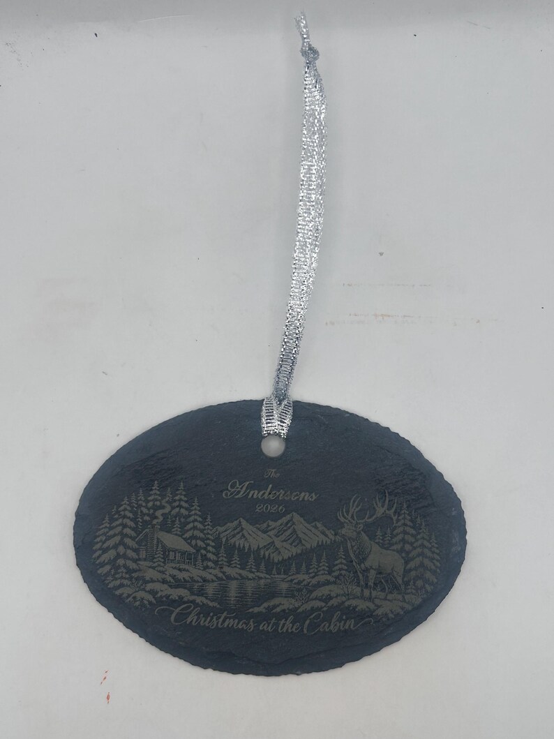 May include: An oval-shaped black slate ornament featuring an engraved mountain landscape with a cabin, a deer, and pine trees. The text reads The Andersons 2026 and Christmas at the Cabin. A silver ribbon is attached through a hole at the top.