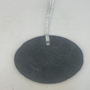 May include: An oval-shaped black slate ornament with a rough, textured surface. The piece features a single circular hole at the top centre, through which a thin, shimmering silver ribbon is looped for hanging purposes.