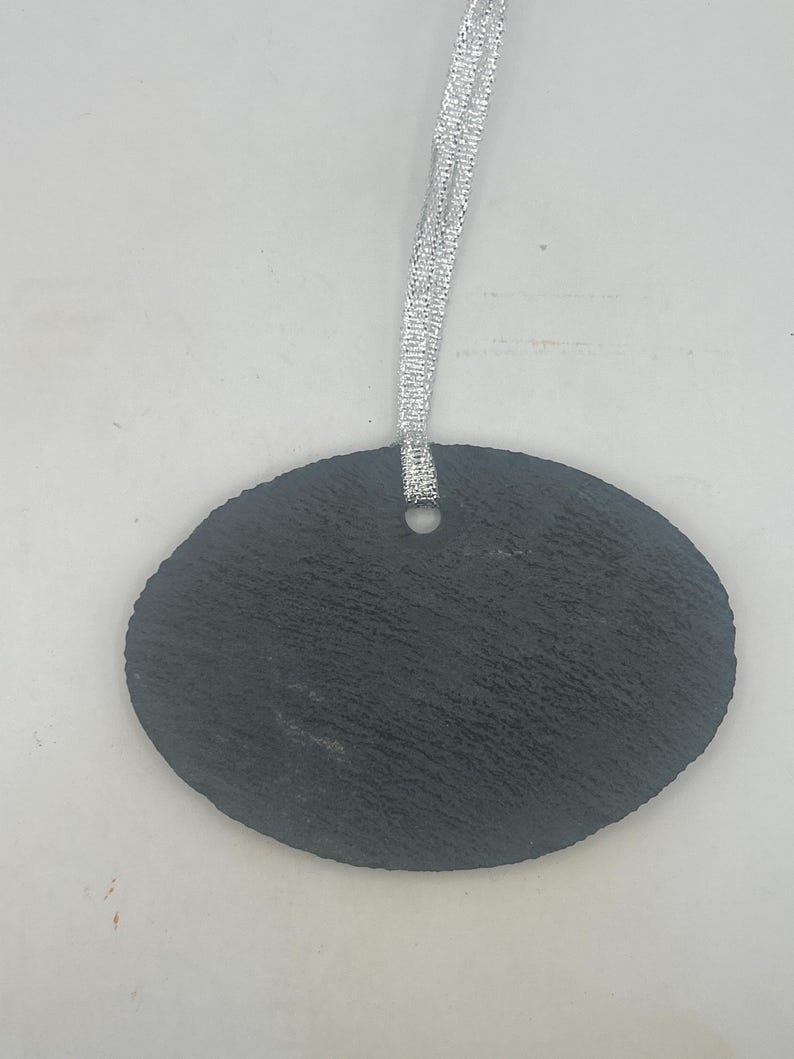 May include: An oval-shaped black slate ornament with a rough, textured surface. The piece features a single circular hole at the top centre, through which a thin, shimmering silver ribbon is looped for hanging purposes.