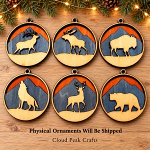 May include: Six circular wooden ornaments featuring laser-cut silhouettes of a deer, moose, bison, wolf, elk, and bear. Each ornament displays a layered mountain landscape in orange and blue tones. Text at the bottom reads: Physical Ornaments Will Be Shipped Cloud Peak Crafts.