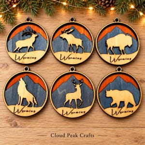 May include: Six circular wooden ornaments featuring laser-cut silhouettes of a deer, moose, bison, wolf, elk, and bear. Each ornament displays a blue mountain range against an orange sky with the word Wyoming printed in black script.