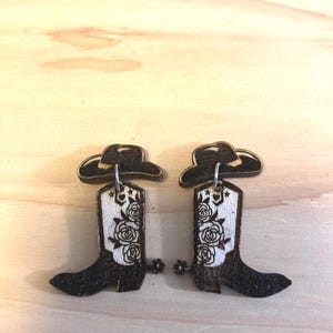 May include: A pair of wooden earrings featuring a cowboy boot design with floral accents and a cowboy hat on top. The boots are black with white floral details and a silver spur.