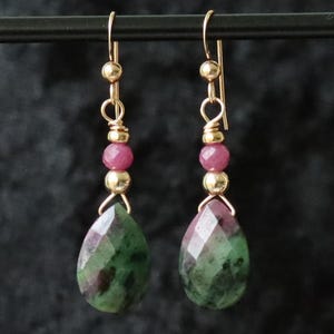 Puede incluir: Pendientes de oro con gotas de piedras preciosas verdes y rosas. Los pendientes tienen una pequeña perla rosa y una perla dorada sobre la gota de piedra preciosa.
