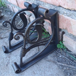 Op de afbeelding: Twee zwarte metalen beugels met ingewikkelde krulontwerpen. De beugels zijn bevestigd aan een bakstenen muur en hebben een decoratieve, sierlijke stijl.