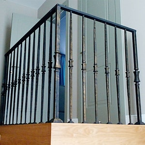 May include: A black metal stair railing with decorative vertical bars and a bronze-colored finish on the decorative elements. The railing is installed on a wooden staircase with light-colored wood steps.