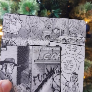 May include: A black and white comic book wallet with panels from a graphic novel. The panels show scenes with a truck, a cafe sign, and characters in a dramatic setting. The text includes phrases like "No, son... this man is alive."