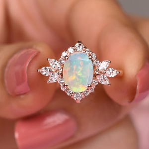 Puede incluir: Anillo de oro rosa con una piedra preciosa de ópalo ovalada rodeada de pequeñas piedras claras. El ópalo tiene un aspecto lechoso e iridiscente. El engaste del anillo tiene un diseño detallado en forma de flor. El anillo se sostiene en una mano con esmalte de uñas rosa.