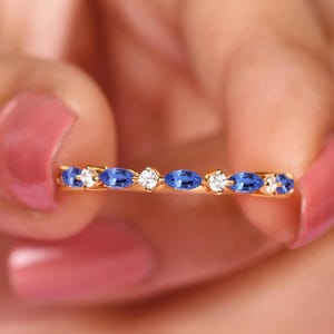 Puede incluir: Un anillo de oro con piedras preciosas azules y claras alternas engarzadas a lo largo de la banda. El anillo se sostiene entre dos dedos con esmalte de uñas rosa. Las piedras preciosas están dispuestas en una fila.