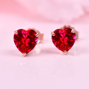 以下が含まれることがあります： 金色の枠にセットされたハート型のルビーのイヤリングのペア。イヤリングは白い背景の上にあります。