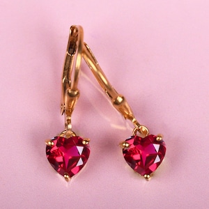 May include: A pair of gold-colored hoop earrings with heart-shaped, ruby-red gemstones. The earrings are displayed on a light pink background. The heart-shaped gems are faceted, catching the light.