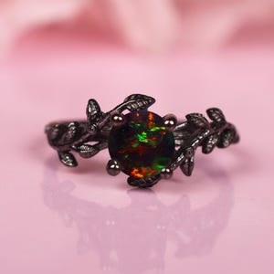 以下が含まれることがあります： 大きな丸い虹色のオパール宝石が付いた黒い指輪。指輪は、宝石を囲む葉が特徴的な葉のデザインです。オパールは、緑、赤、オレンジなど、鮮やかな色の配列を表示します。
