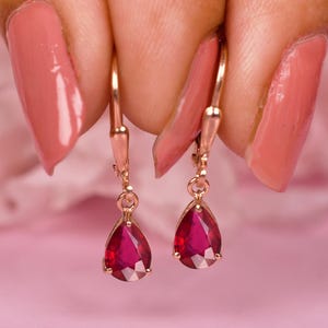 May include: Rose gold drop earrings featuring teardrop-shaped, deep red gemstones. The earrings are displayed against a soft pink backdrop, highlighting their colour and design. A classic jewellery item.