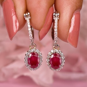 May include: A pair of drop earrings featuring oval-cut deep red rubies surrounded by a halo of small white diamonds. The earrings are suspended from hinged hoops that are also encrusted with tiny white diamonds, set in a polished silver-toned metal.