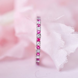 May include: A delicate eternity band with alternating pink and white gemstones. The band is set in a silver metal.
