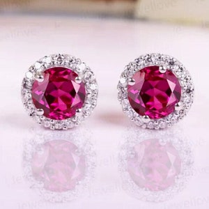 May include: A pair of silver stud earrings with a round, bright pink ruby gemstone surrounded by a halo of smaller white gemstones.