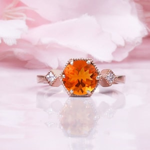 Könnte beinhalten: Ein Ring aus Roségold mit einem großen orangefarbenen Edelstein, der von kleineren klaren Edelsteinen umgeben ist.