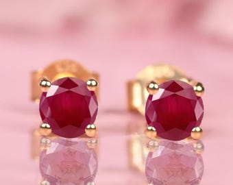 Puede incluir: Un par de pendientes de oro con un brillante rubí redondo de color rojo intenso. Los pendientes se presentan sobre un fondo rosa suave, resaltando el color y el brillo de las piedras preciosas. Joyería clásica.
