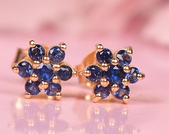 May include: A pair of gold-colored stud earrings, each designed as a flower with six dark blue gemstones. The earrings are set against a soft pink background, reflecting the light.
