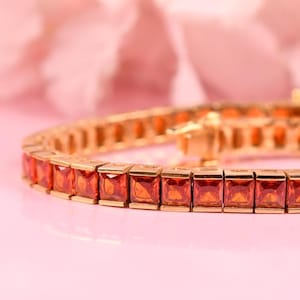 May include: A gold-toned bracelet with square-cut, orange gemstones. The bracelet is set against a soft pink background, with a blurred floral element in the background. The gemstones are evenly spaced and reflect light.