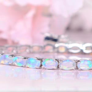 May include: A silver-toned tennis bracelet featuring a continuous row of oval-shaped, iridescent white opals. Each opal is set in a metal prong setting, and the bracelet includes a secure box clasp closure with a safety latch mechanism.