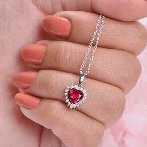 Puede incluir: Un collar de plata con un colgante en forma de corazón. El colgante presenta una gema roja vibrante en el centro, rodeada por un halo de gemas más pequeñas y claras. El collar se sostiene en una mano con esmalte de uñas color coral.