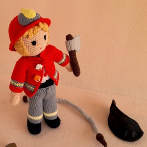 May include: A crocheted firefighter doll with a red jacket, gray pants, and a yellow helmet. The doll is holding a small brown axe and has a gray hose attached to its back.