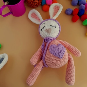 Könnte beinhalten: Ein rosa gehäkeltes Häschen mit einem lila Herz auf der Brust und weißen gehäkelten Ohren. Das Häschen trägt einen lila gehäkelten Schal um den Hals.