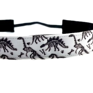 May include: White headband featuring a repeating pattern of black dinosaur skeletons and bones. The headband has a black elastic band for a secure fit. This accessory is perfect for adding a touch of prehistoric flair to any outfit.