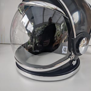 May include: A black and silver astronaut helmet with a clear, curved visor. The helmet has a reflective surface, with a silver trim and a white cord. The visor reflects the surrounding environment.